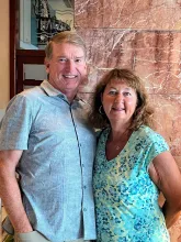 A man and a woman standing close together and smiling at the camera. The man has short, reddish-blonde hair and is wearing a light blue, short-sleeved button-down shirt. The woman has wavy, shoulder-length brown hair and is wearing a turquoise v-neck top with a blue and green floral pattern. They are indoors, standing in front of a textured, reddish-brown marble wall.